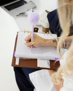 Person writing in a planner with a decorative pen, surrounded by office supplies.